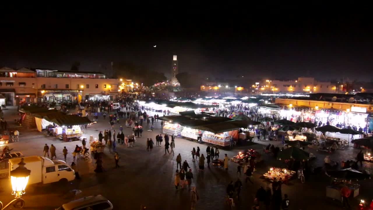 amplia vista aérea del mercado nocturno de marrakech, marruecos, con turistas y lugareños explorando los zocos y puestos de vendedores de comida y recuerdos con la luna creciente elevándose sobre la mezquita.