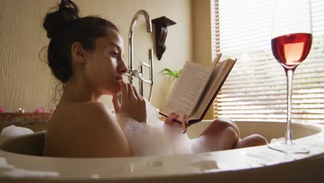 relaxed biracial woman lying in bath with foam and glass of wine, reading book