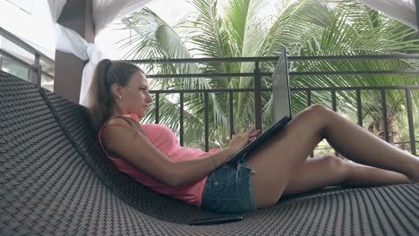 girl lies on curved sunbed and types on laptop keyboard