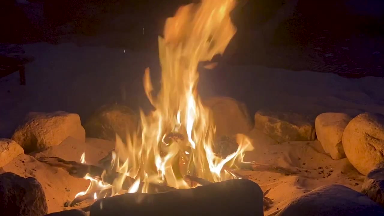 Un falò vivace con fiamme arancioni e gialle brillanti che brucia in un braciere circolare in pietra posizionato nella sabbia di notte, creando un'atmosfera calda e accogliente