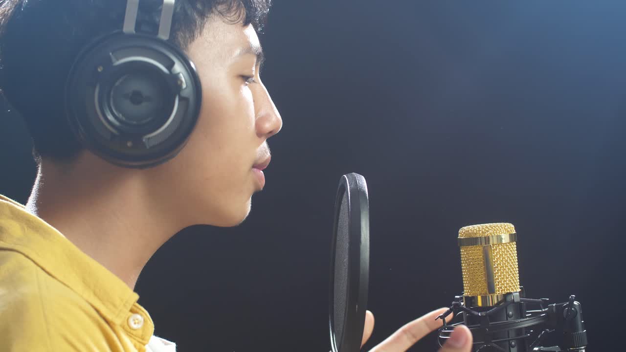 흑색 배경 에 있는 콘덴서 마이크 로 노래 하는 헤드폰 을 착용 한 젊은 아시아 소년 의 근접적 인 측면 시각