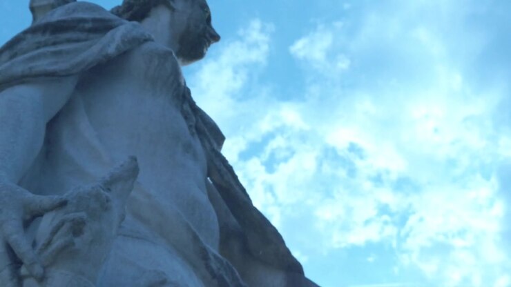 Camera tilts up along a carved marble statue with detailed drapery against a bright sky. Sunlight highlights the sculpture while clouds create a soft background in Rome Italy