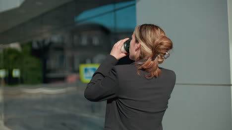 side view of professional woman sipping coffee with hair tied back on phone call as she walks by glass building reflecting surroundings in bright urban setting with blurred background and sunlight