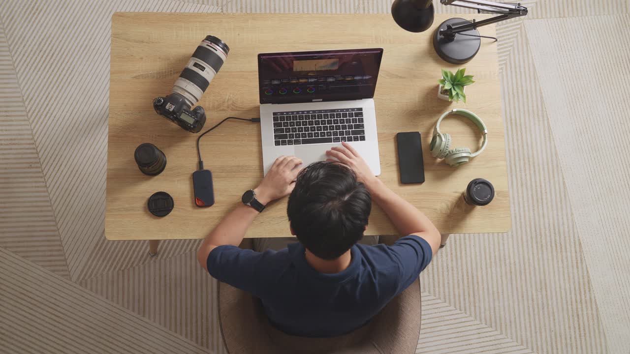 Top View Of A Male Color Grading Sitting In The Workspace Using A Laptop Next To The Camera Editing The Video At Home