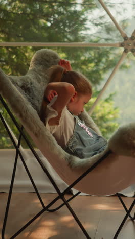 little girl basking in cool fur chair and getting comfortable for afternoon nap. child watches landscapes of forest and mountains behind transparent window of glamping