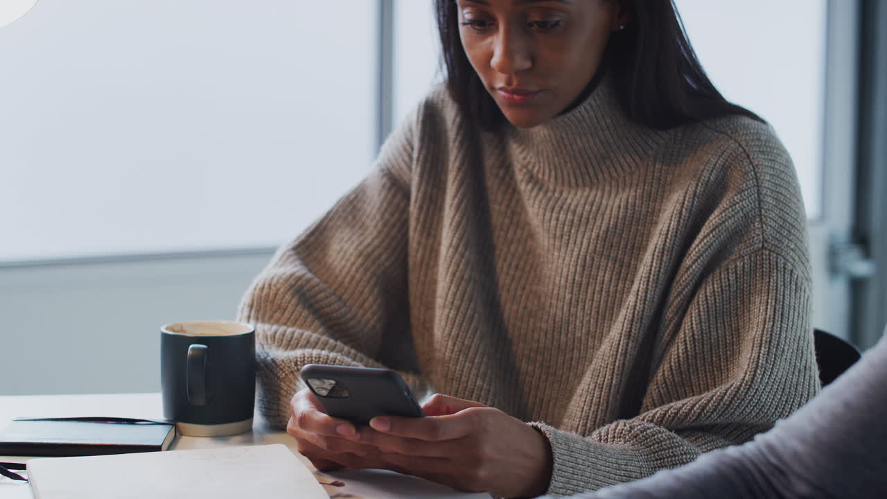 Businesswoman Using Mobile Phone As Male Colleague Works On Laptop In Foreground