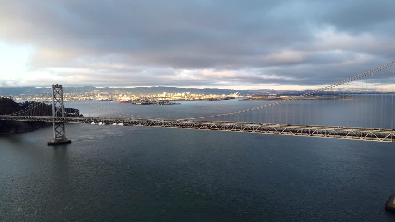 샌프란시스코 오클랜드 베이 브리지 더블 서스펜션의 4k 30fps 공중 드론 영상 - 자동차는 고속도로, 컨테이너선, 폭풍우 치는 구름, 잔잔한 베이 물, 파도에 반사되는 일몰을 따라 이동합니다.