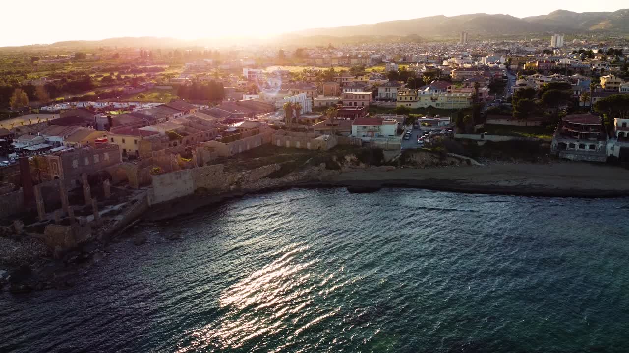 Ripresa aerea con drone all'ora d'oro su Avola, Sicilia. Storica città costiera con antiche fortificazioni lungo il litorale, calda luce del tramonto che inonda il paesaggio urbano e il Mar Mediterraneo sottostante.