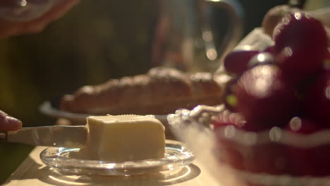 Lifting-butter-dish-on-outdoor-table-to-slice-and-spread-on-toast-in-background