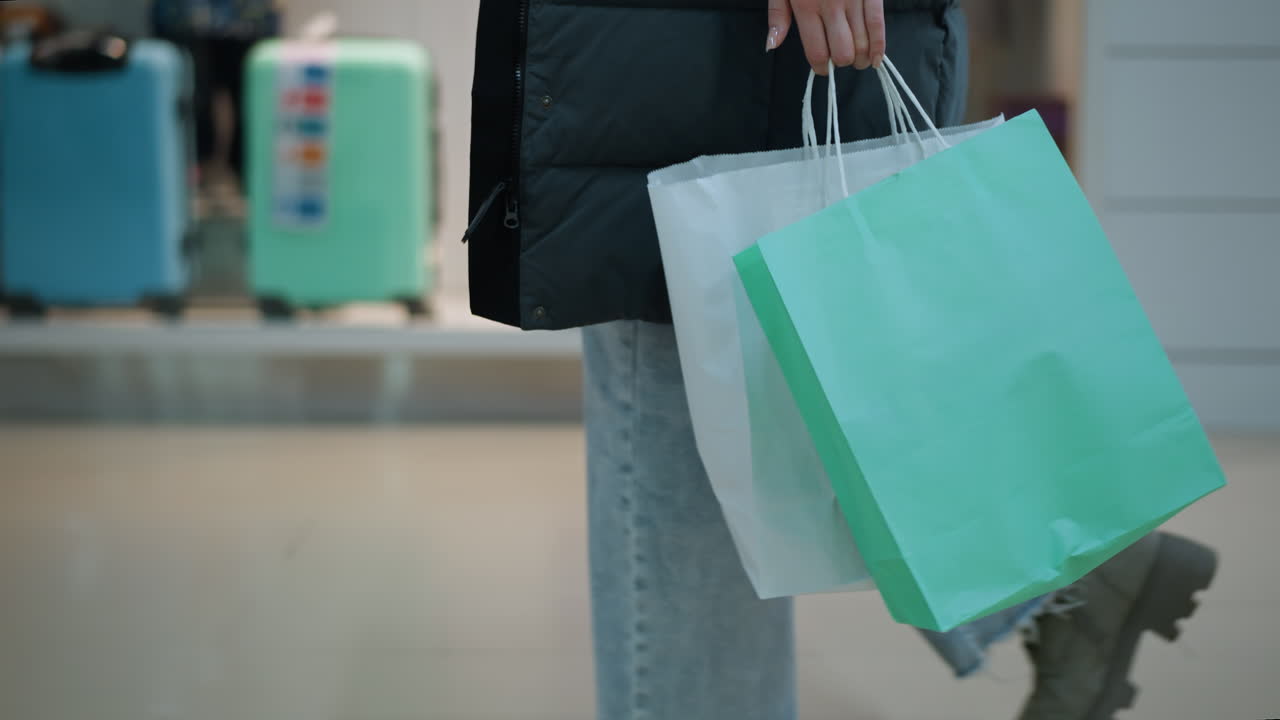 잘 조명 된 쇼핑백, 옷을 입은 마네킨 및 유리 디스플레이 내에 배열 된 다채로운 여행 가방을 들고 잘 조명된 쇼핑 공간을 지나가는 사람의 측면 시각