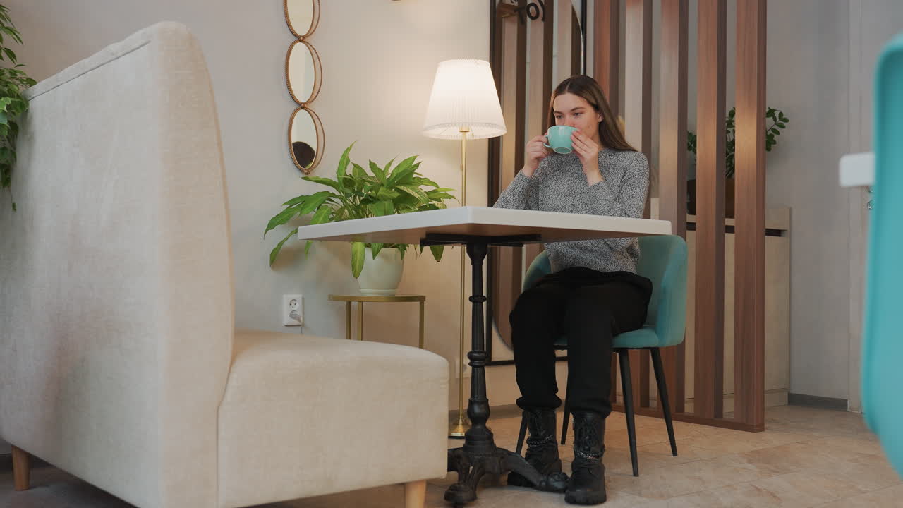 Young woman seated enjoying cold drink with deep relief on face in cozy indoor space featuring lamp stand, decorative plant, phone on table, empty chair opposite her, and door in background