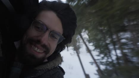 Close-up-of-young-man-face-with-beard-and-long-hair-and-glasses-smiling-enjoying-his-train-ride-through-snowy-natural-landscape,-sustainable-green-travel-concept-hanging-outside-window