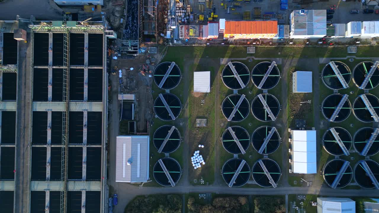 Industrial complex with a chimney emitting smoke into the sky, next to a sewage treatment facility and urban infrastructure. Smooth aerial view vertical bird's eye view drone camera pointing down