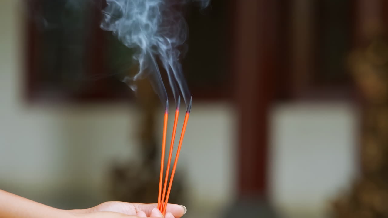 Manos asiáticas sosteniendo varitas de incienso encendidas, tres varitas naranjas emiten humo ondulante, fondo de templo borroso