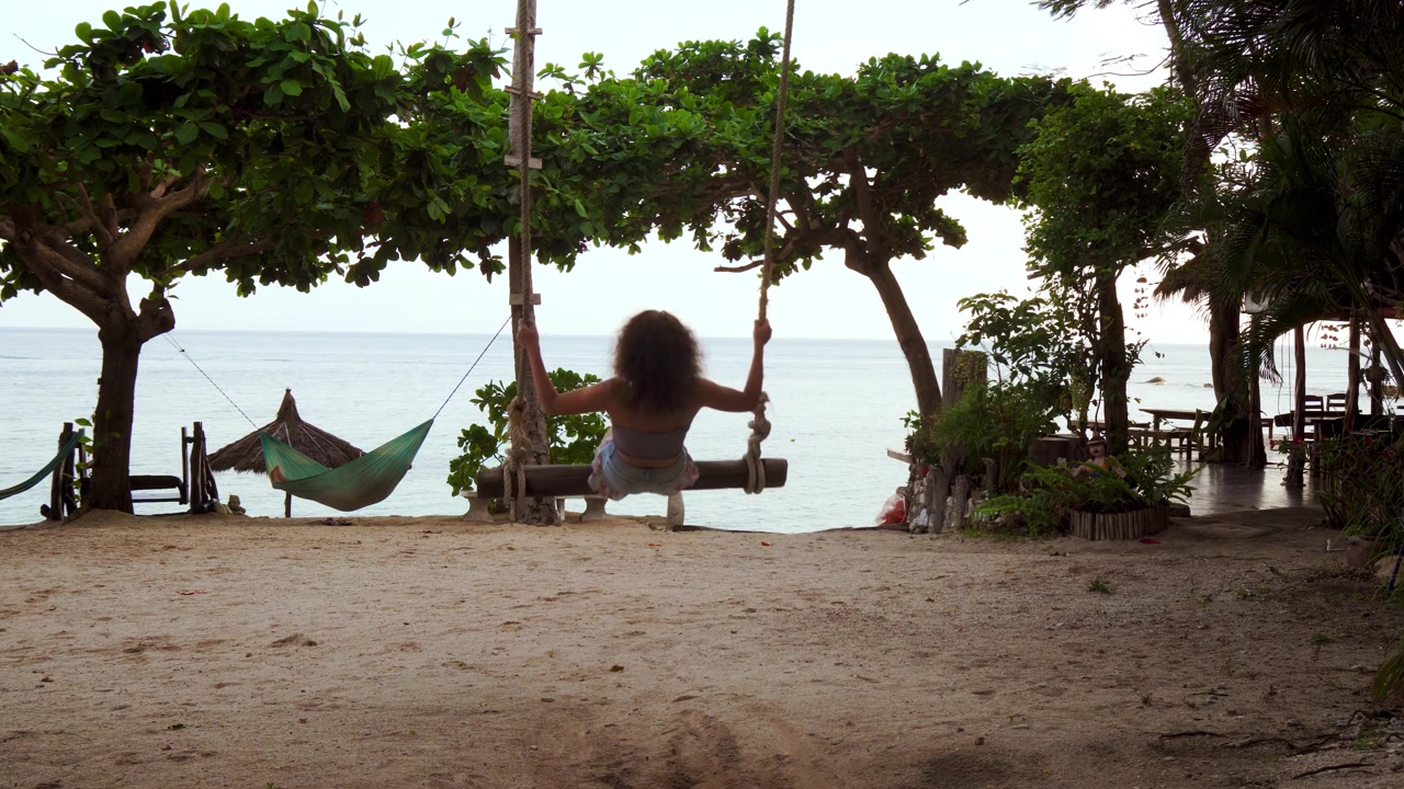 Giovane donna che si dondola verso l'orizzonte dell'oceano su un'altalena di corda, amaca e alberi tropicali, Koh Tao, Thailandia, telecamera fissa