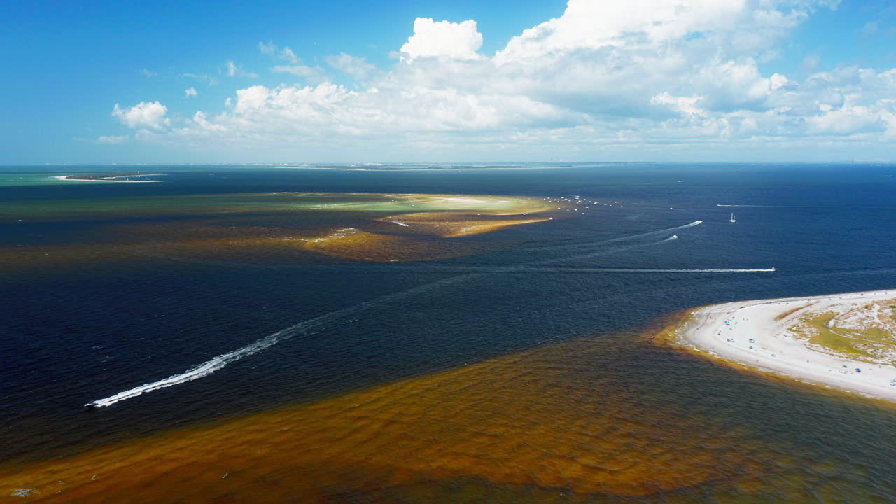 Boats carve bright white wakes across emerald and deep blue waters beside a sandy peninsula, where shifting sandbars create dramatic color contrasts in the expansive seascape