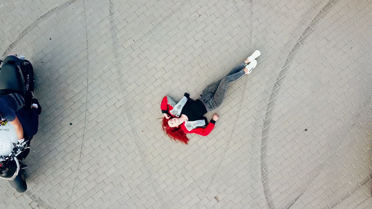 Close-up of an extreme red-haired woman is lying on the road outdoors. Beautiful young female and three motorbikers riding around her. Aerial view. Camera moves up around in a circle.