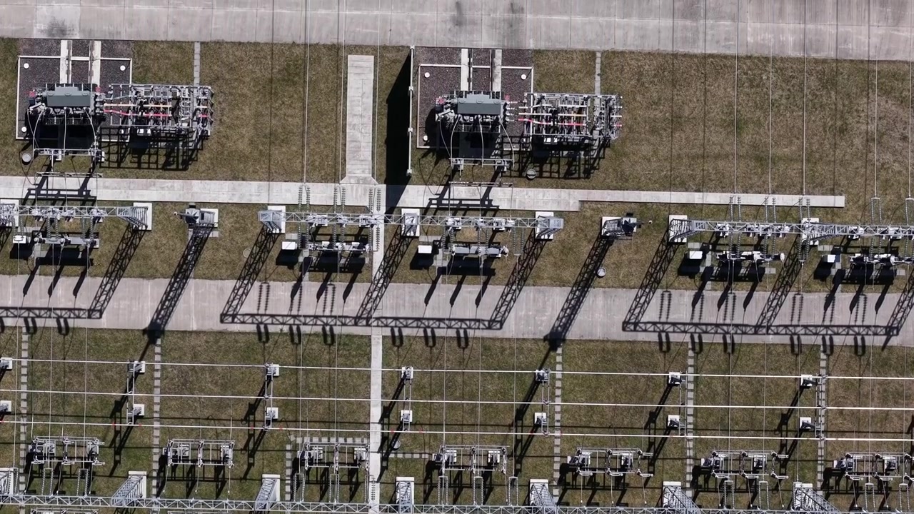 Letecký pohled z vysokého úhlu ukazuje komplexní infrastrukturu elektrické rozvodny s vysokonapěťovým zařízením a dlouhými stíny na travnatém poli.