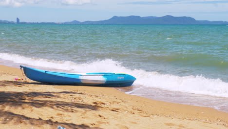 A-blue-and-white-kayak-on-the-beach-as-waves-rush-to-the-shore,-a-lovely-horizon-with-mountains-during-a-warm-and-perfect-day