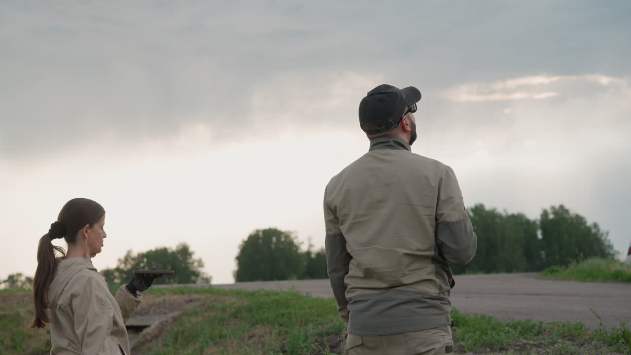woman in protective suit releases black balloon using handheld device while standing beside male colleague in open grassy field under overcast sky