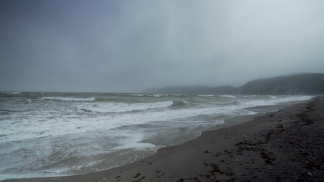 waves roll in creating sea foam on an empty beach with fog covered sea cliffs in the background on a depressing cold wintery morning, perce quebec