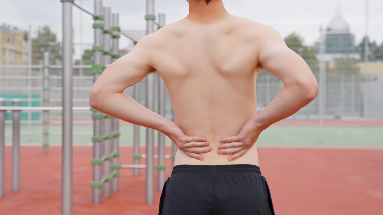 A young athletic man rubs his lower back in pain while exercising outdoors in a sports park