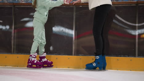 a close-up view of a child and her parent playing on an ice rink, the child is wearing mint green cloth and pink ice skates, they are standing near the edge of the rink