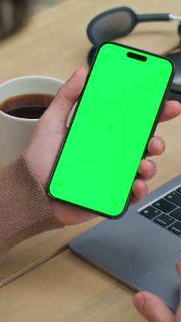 person using a smartphone with green screen and laptop at a table