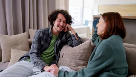 Happy-brunette-guy-with-curly-hair-in-a-gray-checkered-shirt-sits-on-the-sofa-and-communicates-with-a-brunette-girl-while-spending-time-together-in-a-modern-apartment