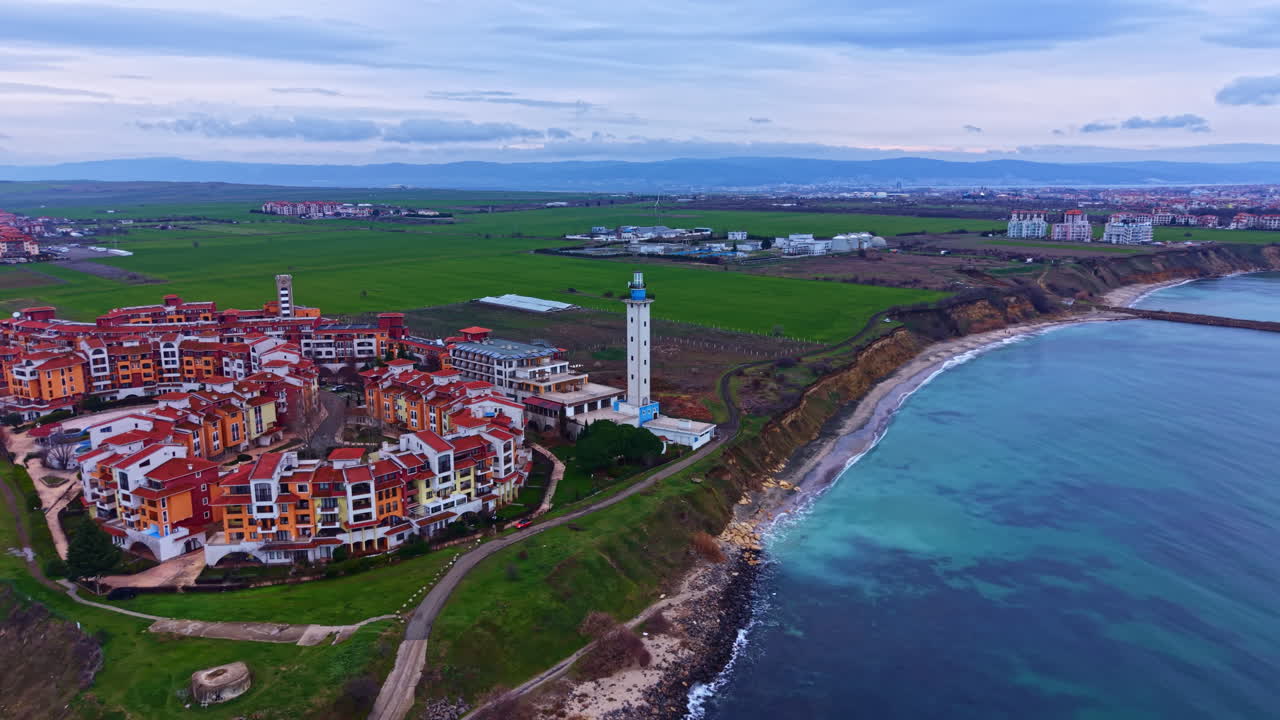 Coastal view of a small town with houses, a lighthouse, and green fields in the background under a cloudy sky