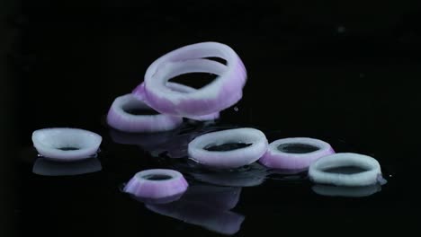 disparo invertido en cámara lenta de anillos de cebolla blancos y púrpura que caen sobre una superficie negra una superficie cubierta de agua