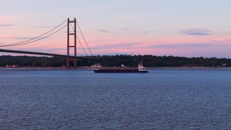 witness the serene passage of a barge boat beneath the humber bridge at sunset in this captivating drone video