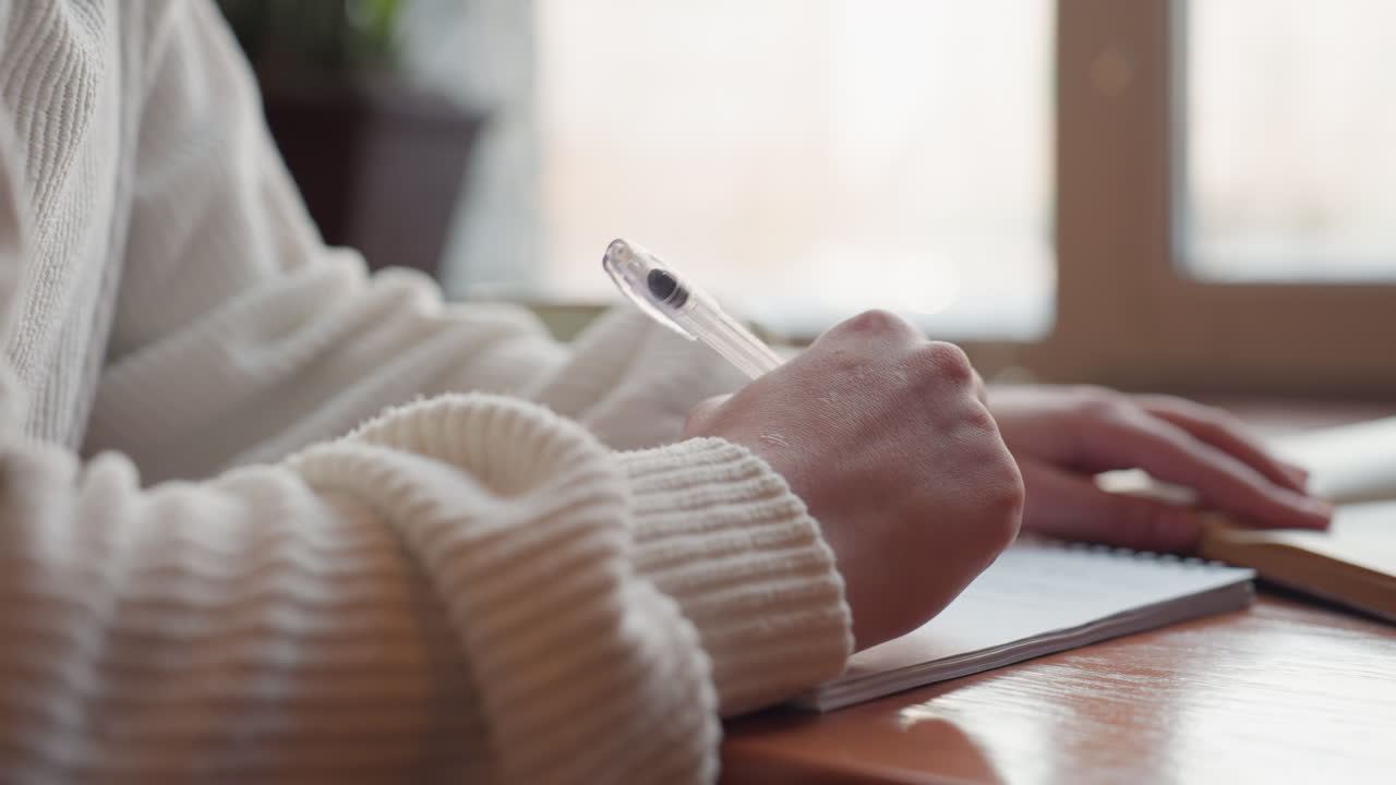 close up of lady hand holding transparent pen writing in notebook on wooden desk beside open book with natural light from window creating calm productive atmosphere for focused work