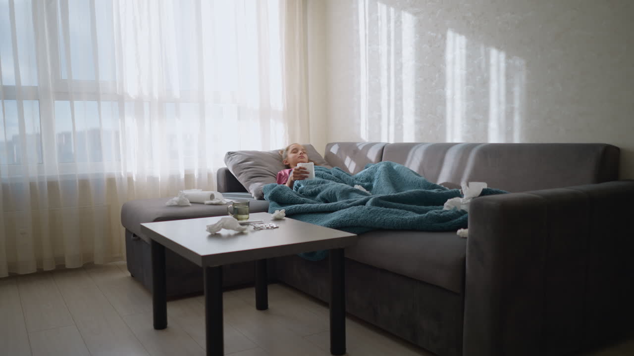 Unwell child lying on couch with tissue paper scattered around her, sick child covered in blanket resting in living room, surrounded by tissues, appearing tired and sick