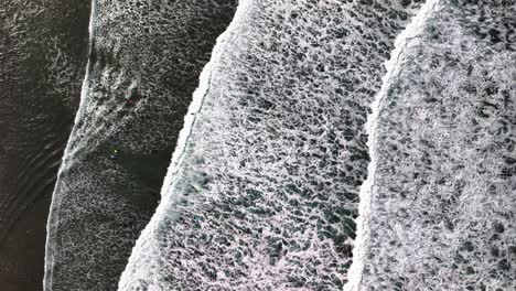 High aerial shot of large, powerful, waves with reflective, large white caps, crashing over a scenic Cornish beach and over the wating surfers