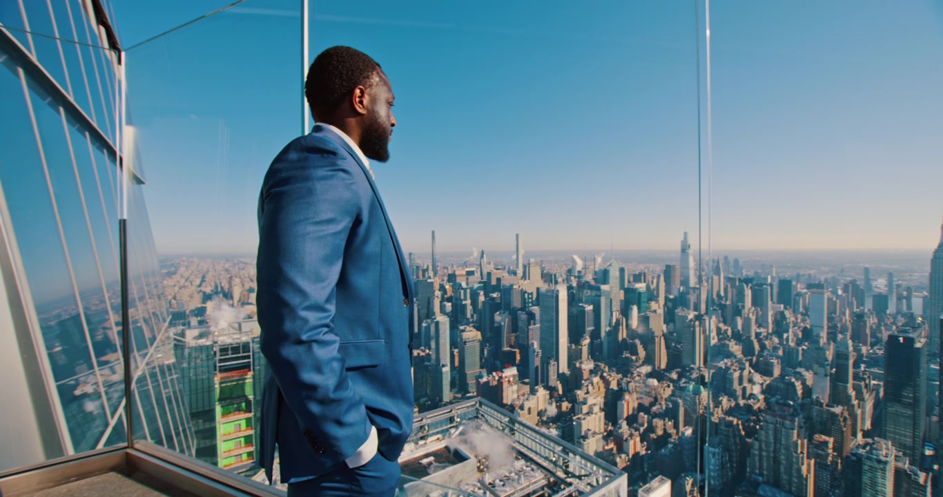 New York, USA, 1 May 2024: Side profile of man in blue suit gazing over New York City. Close framing from glass platform with dense urban towers in warm daylight.