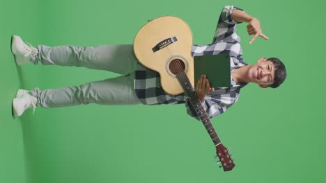 full body of asian teenager with guitar pointing to a tablet in hand while standing on green screen background
