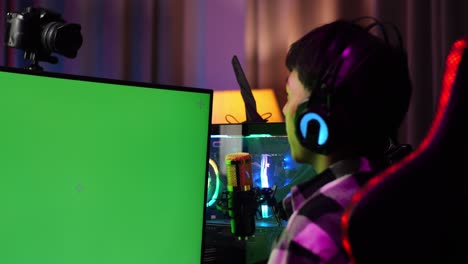 close up back view of asian boy streamer using green screen personal computer, live stream waving hand and talking on the microphone.  desk illuminated by rgb led strip light