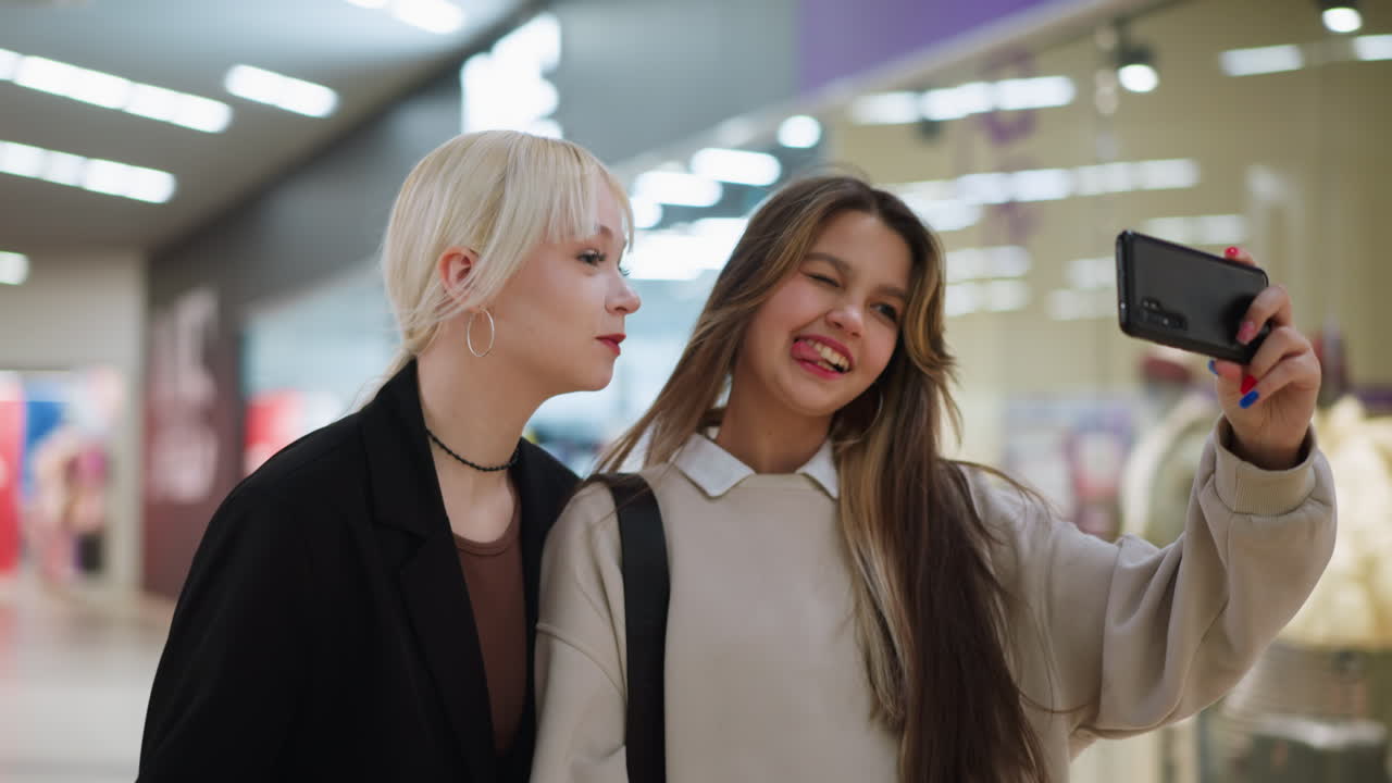 Two young teens take selfie with smartphone while walking past clothing store inside shopping mall, smiling and posing together under bright interior lights with colorful clothes in background
