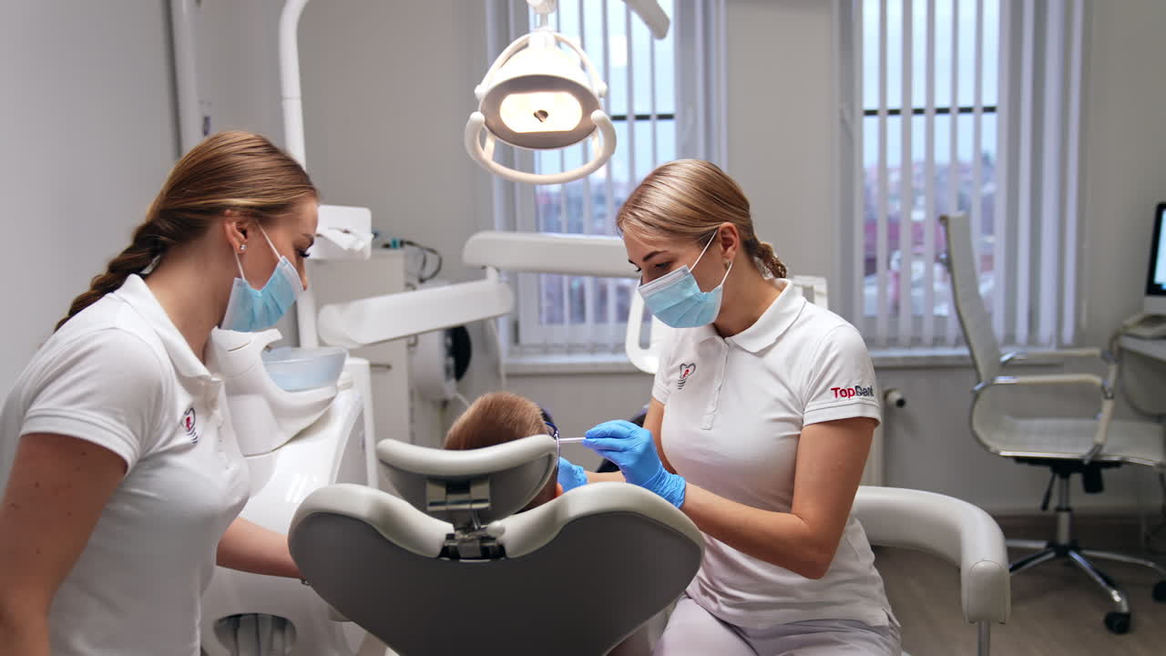 Young beautiful female dentist taking a metal instrument. Doctor is starting dental examination of a little boy. Nurse sitting on the other side of a patient.