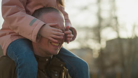 father smiling with daughter playing with his face in the park during sunset, both laughing joyfully