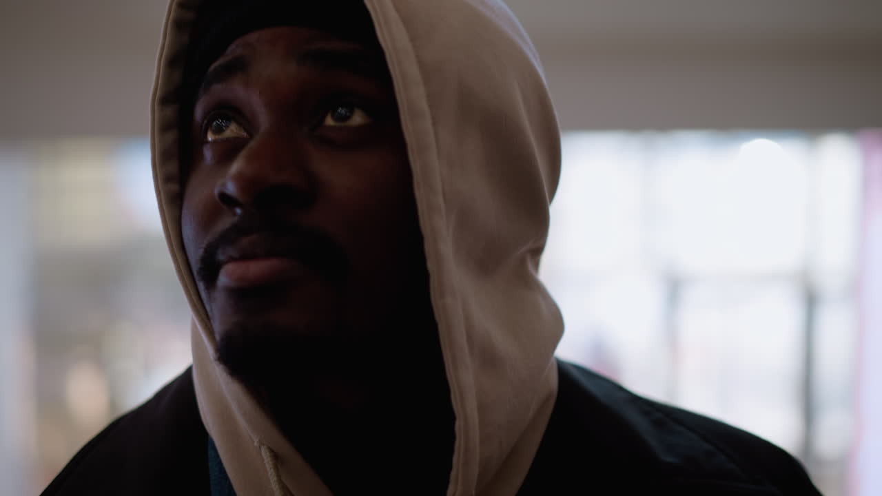 Young black man wearing beige hoodie and black beanie gazing upward in bright indoor space with strong backlight and blurred background capturing contemplative expression and soft ambient light
