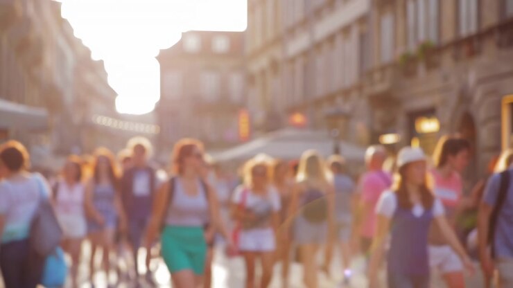 Persone si muovono verso e lontano dal punto di vista attraverso una strada urbana affollata, mentre una forte luce solare calda risplende tra gli edifici, creando una sfocatura morbida, controluce e un leggero bagliore dell'obiettivo