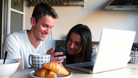 Couple-using-mobile-phone-and-laptop-while-having-breakfast