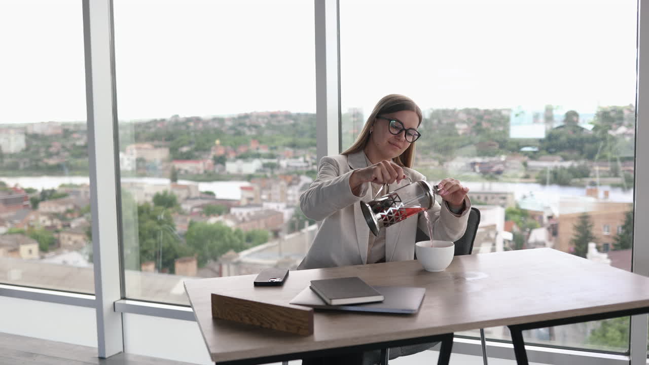Ung kvindelig leder med briller arbejder på et lyst kontor og drikker te ved en bærbar computer. Attraktiv langhåret kvinde med briller på sin arbejdsplads.