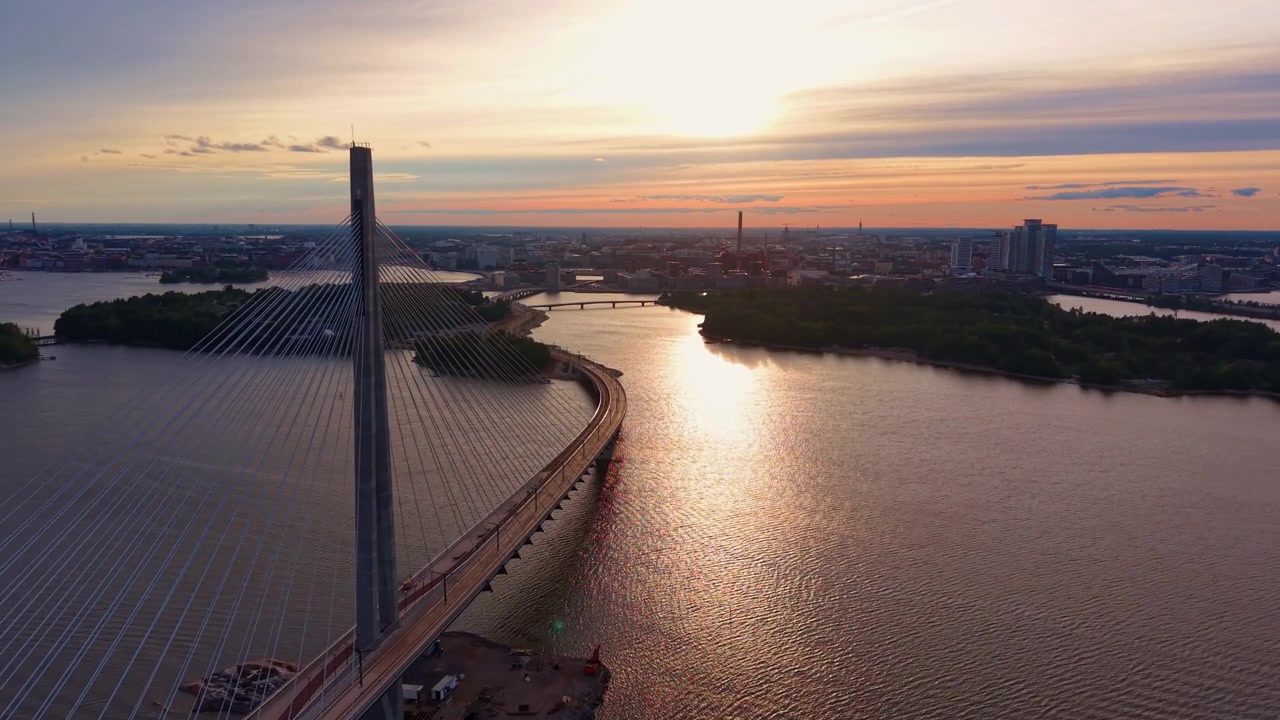 Il ponte strallato Kruunuvuorensilta si curva sulla baia di Helsinki al tramonto, con la luce dorata che si riflette sull'acqua calma e lo skyline della città e isole verdi oltre
