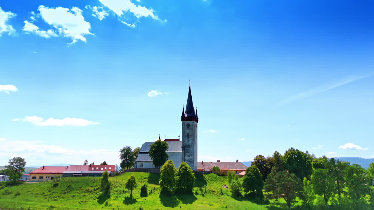 Church on hilltop in summer daylight. A historic church stands on a grassy hill with trees, surrounded by colorful buildings under a bright blue sky