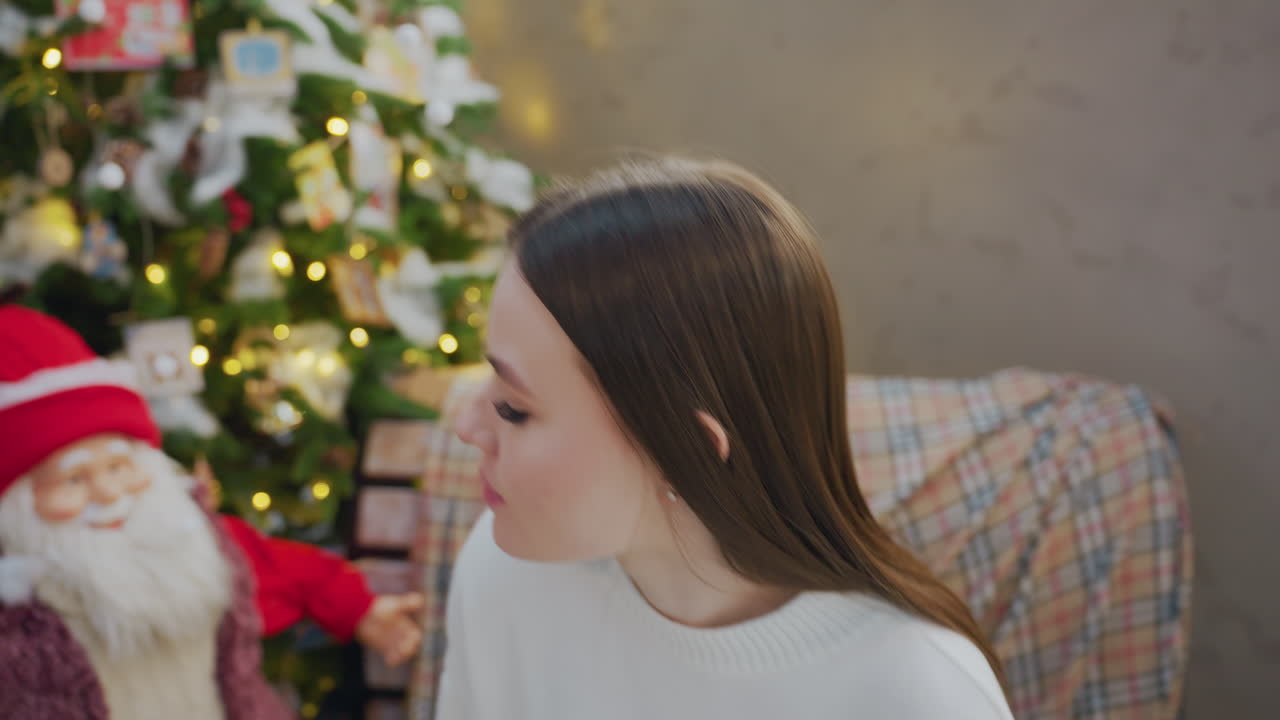 Close-up of woman seated in front of a Christmas tree, gazing at Santa Claus figure by her side, with twinkling lights and holiday decorations in the background
