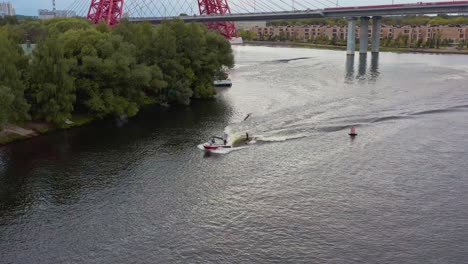 Aerial-following-wakesurfer-and-boat,-bridge-in-the-foreground,-Moscow,-Russia