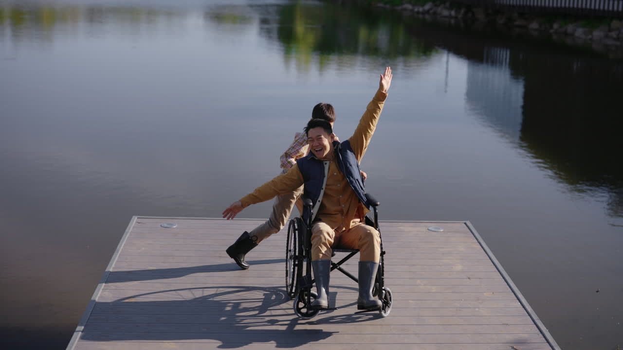Boy Helping Man in Wheelchair on a Dock by a Lake